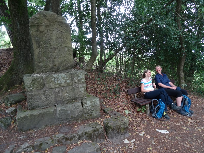 Zwei lachende Frauen mit Rucksäcken auf einer Bank neben einem Denkmal.
