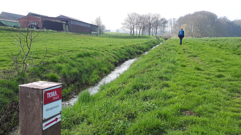 Das Bild zeigt im Vordergrund das Schild des TERRA.track Königsbrück, im Hintergrund Meike und den Bauernhof. Links ist ein kleiner Bach zu sehen.